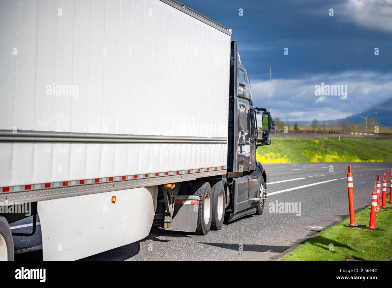 Industrial professional black big rig bonnet semi truck transporting commercial cargo in dry van semi trailer with aerodynamic skirt running on the lo Stock Photo