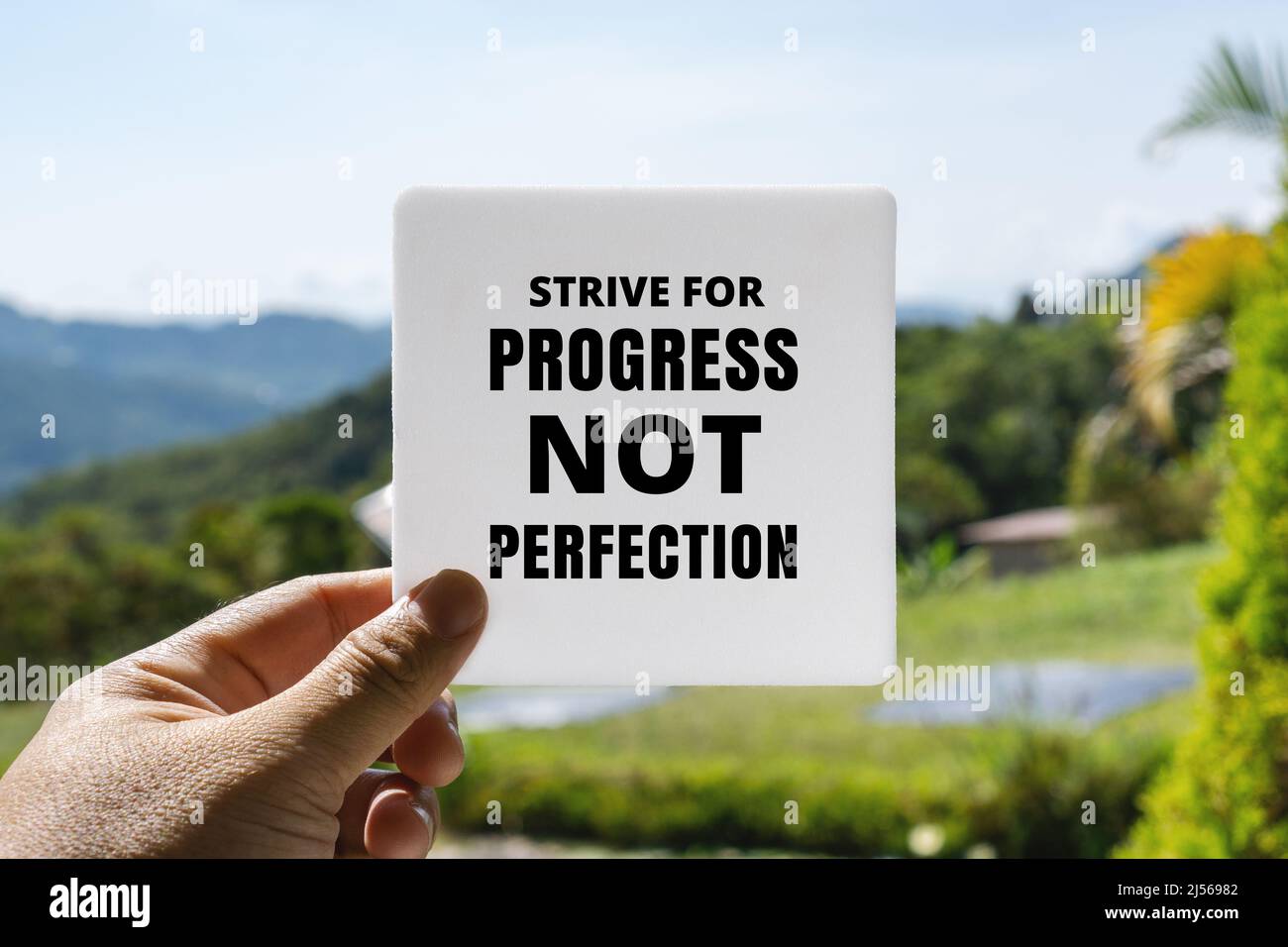 Hand Holding White Paper Card Against Fresh Nature Background. Inspirational Quote. Strive For Progress Not Perfection. Stock Photo