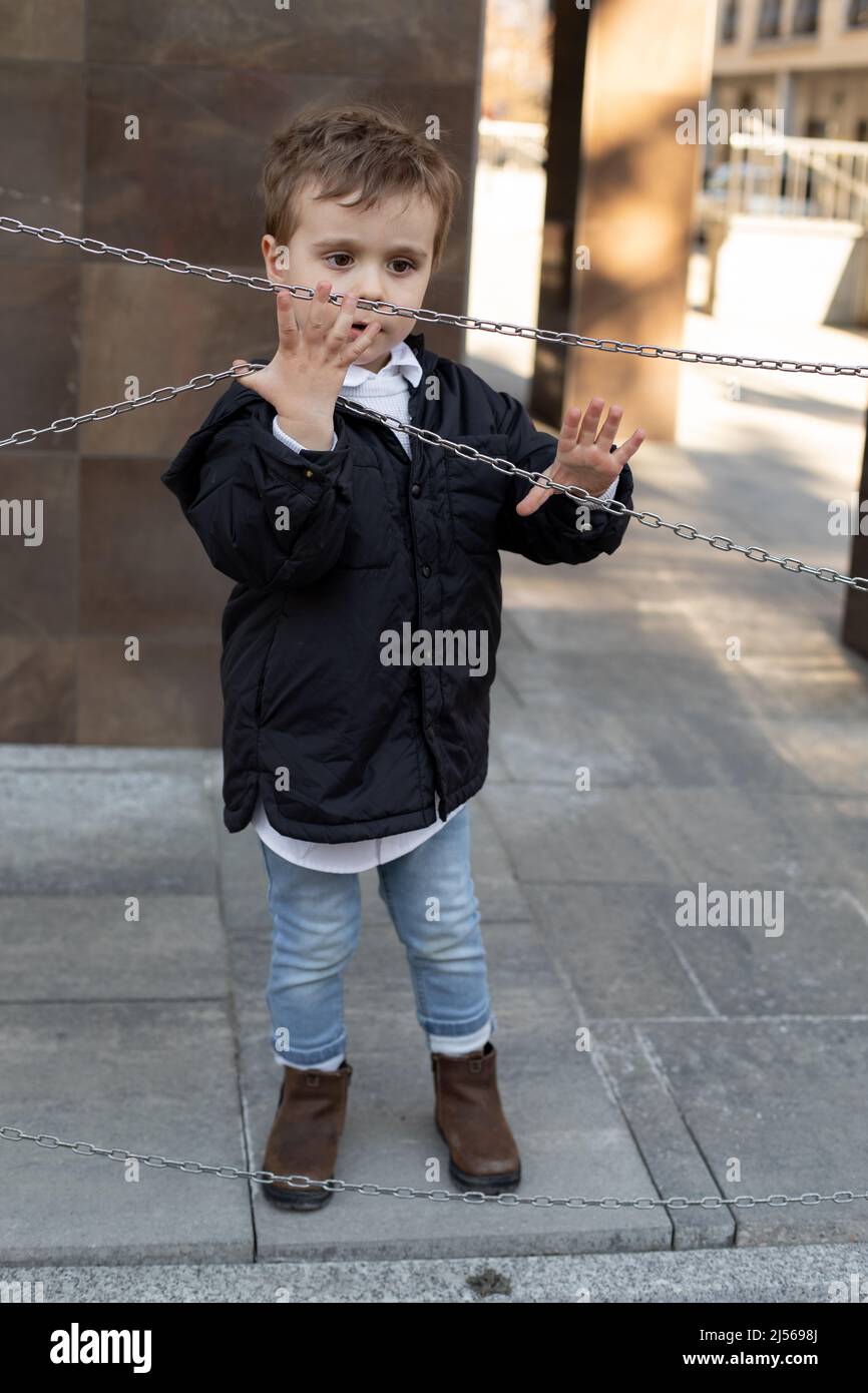 Vertical photo of cute little boy in black jacket standing on the street, holding chains. Developmental abnormalities Stock Photo