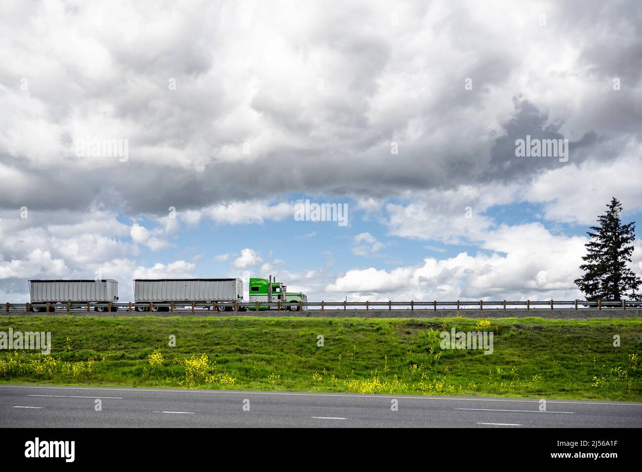 Industrial powerful classic bonnet American green big rig semi truck tractor with two loaded bulk semi trailers transporting cargo running on the road Stock Photo