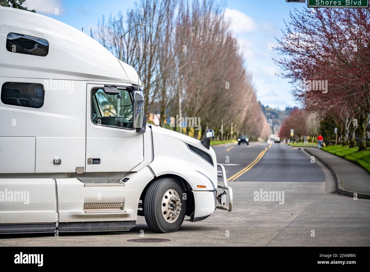 Classic white big rig industrial standard semi truck with dry van semi trailer transporting commercial cargo running on straight local city street roa Stock Photo