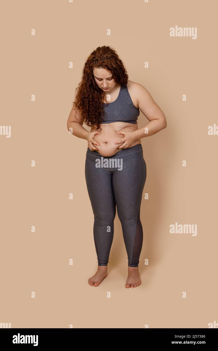 Vertical puzzled red haired overweight fat barefoot woman looking on tummy, doing activity, holding touching belly Stock Photo