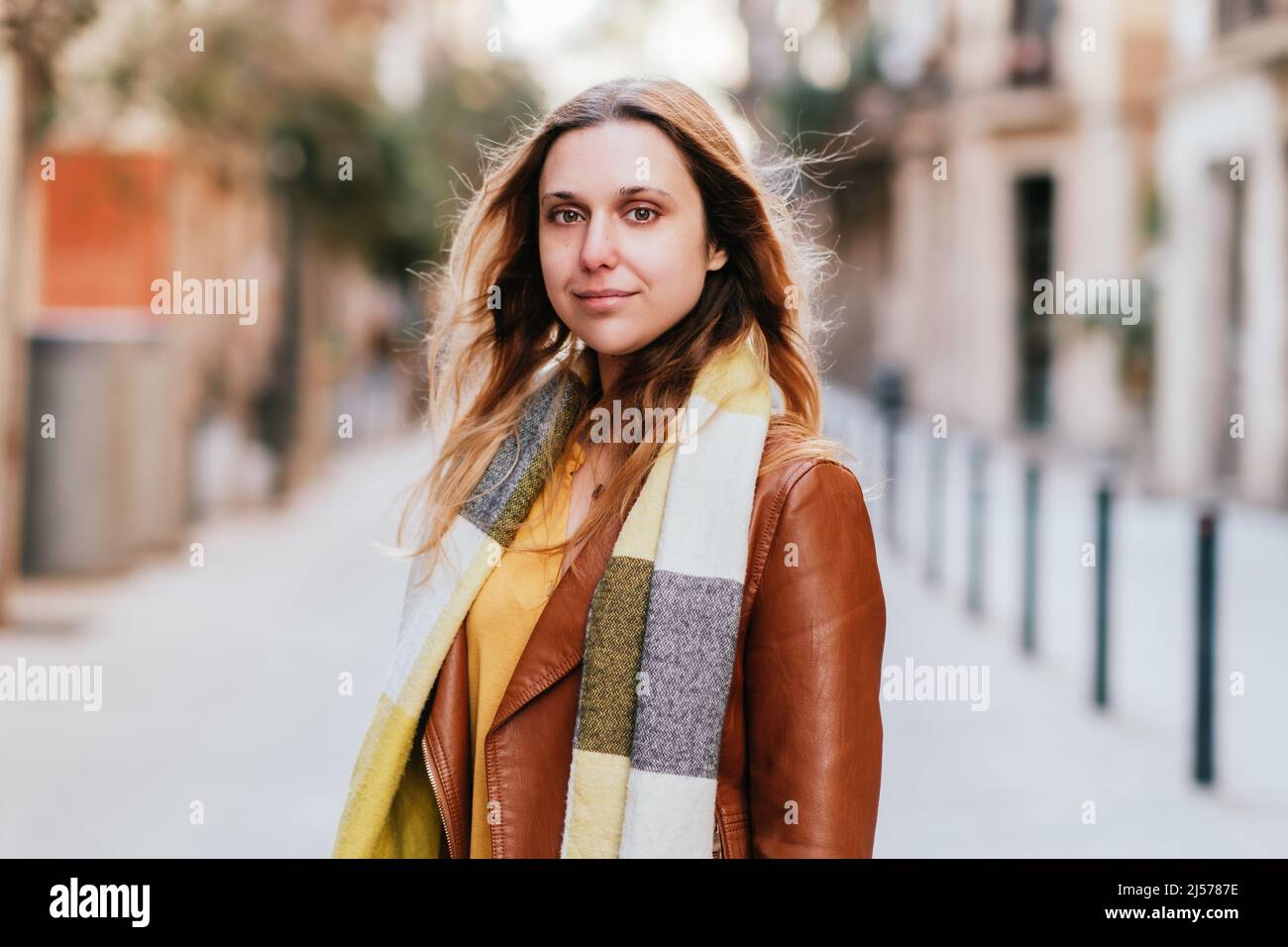 Charming female in leather jacket and scarf looking at camera while standing on sidewalk in city with buildings on blurred background Stock Photo