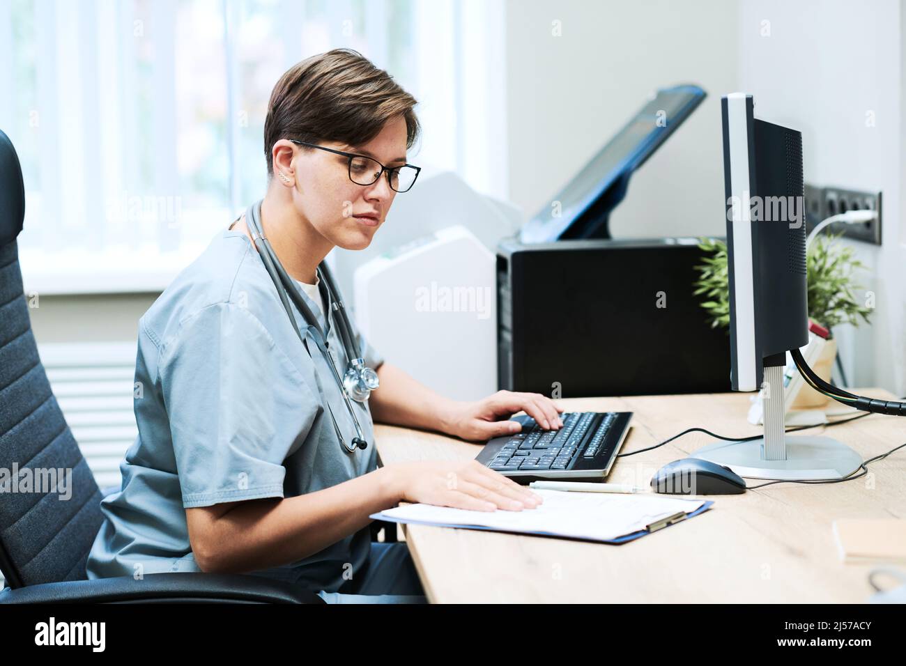 Concentrated young nurse in eyeglasses sitting at desk and using