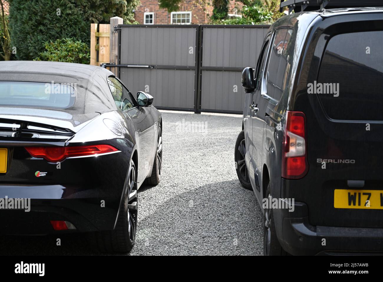 a view behind two parked black vehicles, a sports car and a van, on a grey stone driveway with grey and black entrance gates Stock Photo
