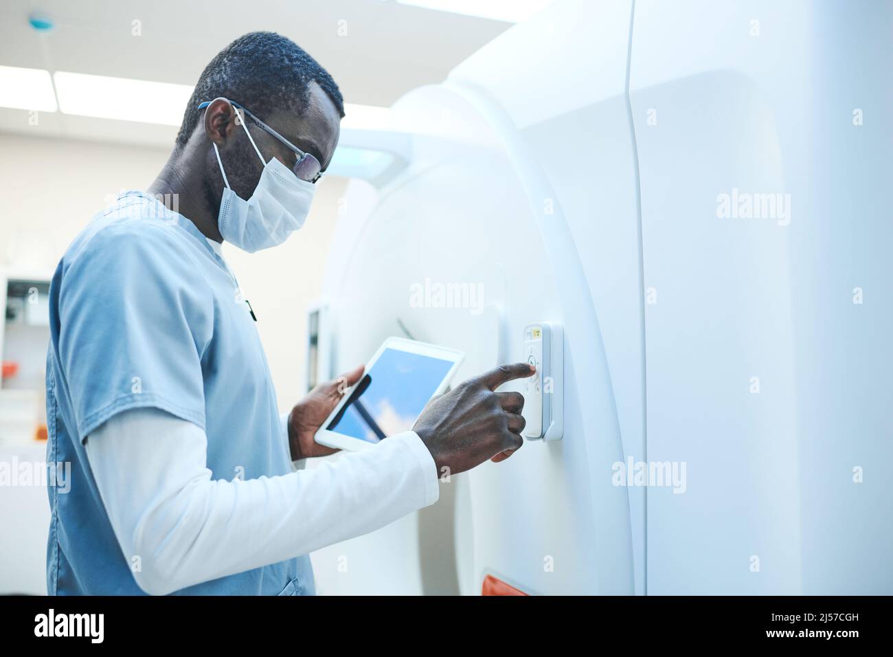 African-American MRI technologist in mask and glasses holding tablet while turning on equipment Stock Photo