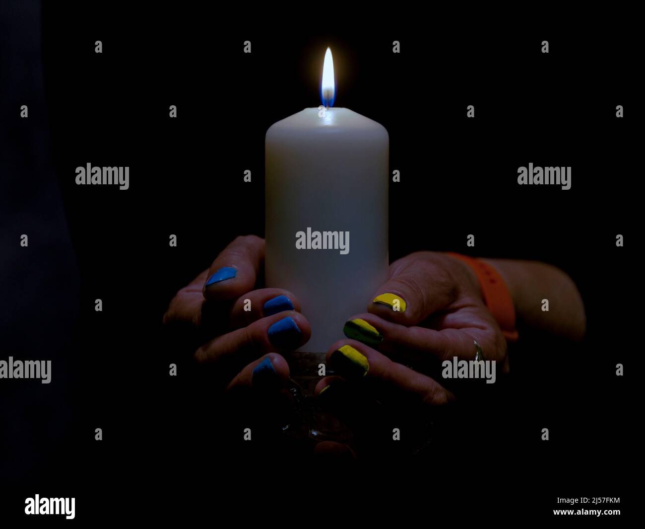 The flame of a white candle in the dark held by a woman. A woman's nails are painted blue and yellow. Stock Photo