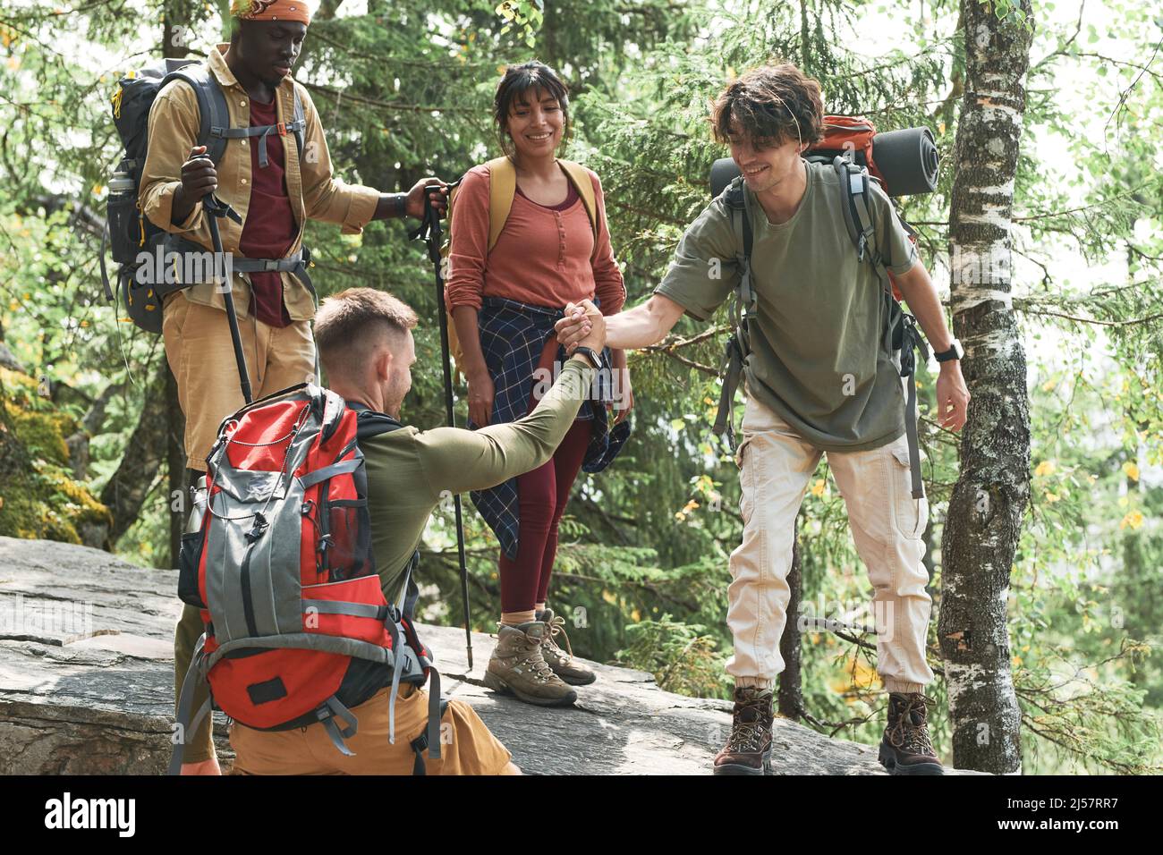 Smiling young man holding hand of friend with backpack tight while assisting him to get up stone at forest hike Stock Photo
