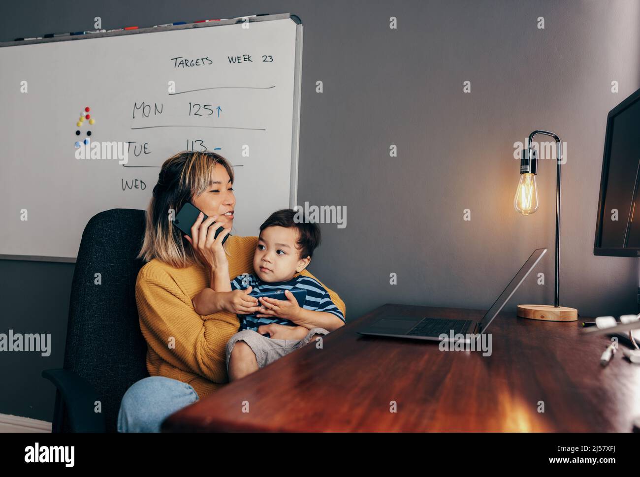 Mother of one working in her home office. Happy working mom speaking on the phone while carrying her son on her lap. Single mom communicating with her Stock Photo