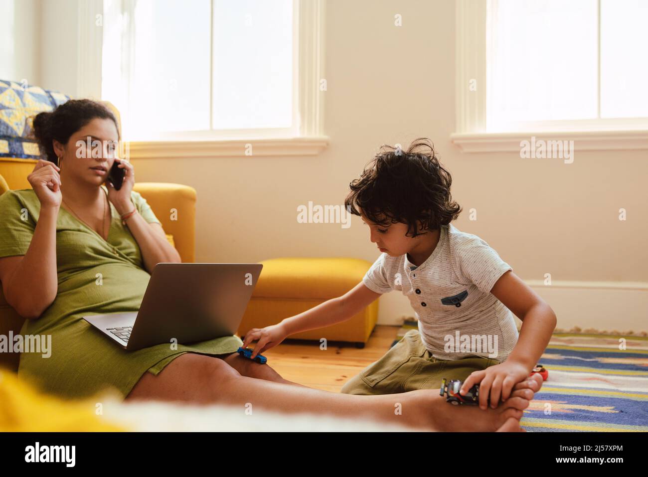 Single mother speaking on the phone while sitting in her son's play area. Working mother making plans with her clients over a phone call. Young self-e Stock Photo