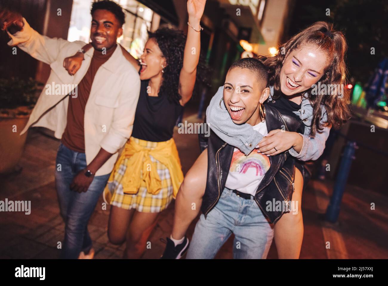 High-spirited young woman piggybacking her best friend at night. Happy young women having fun while going out with their friends in the city. Vibrant Stock Photo