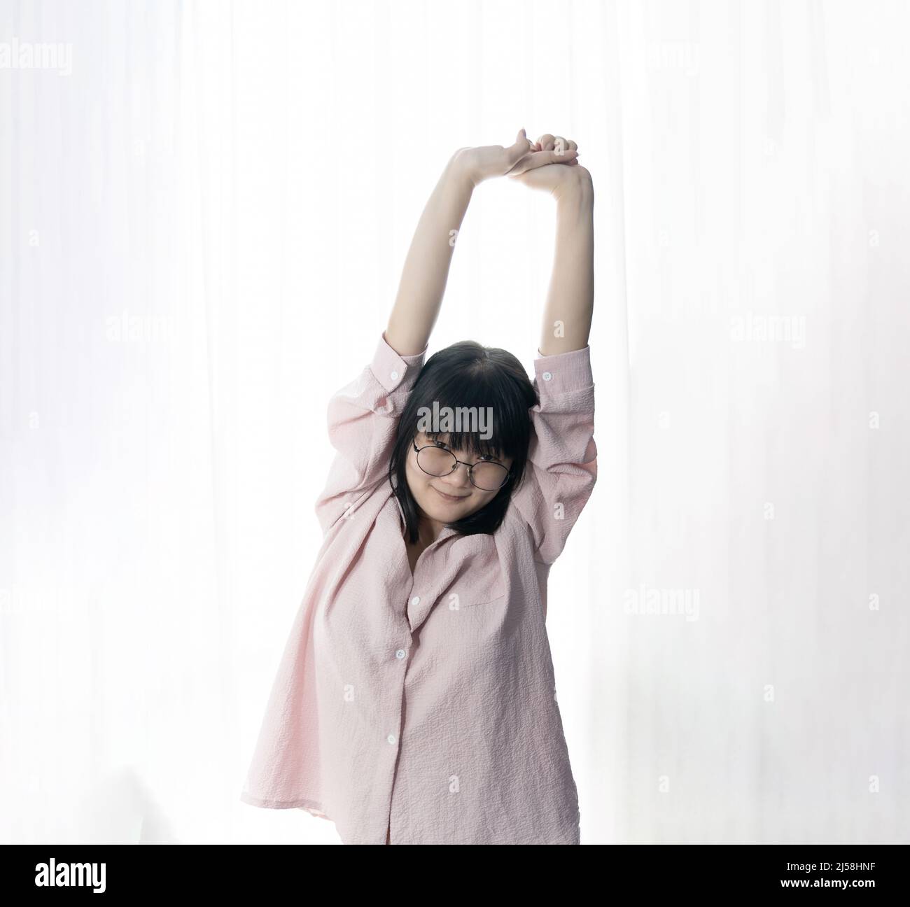 Standed Little Asian young Glasses Girl stretches oneself in front of light white curtain background. Stock Photo