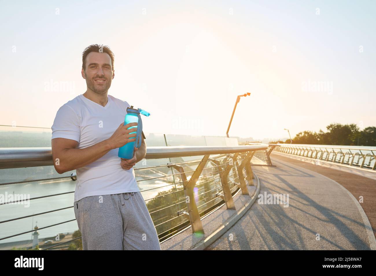 Confident athlete, Caucasian man with muscular aesthetic body in sportswear holds a bottle of water, refreshes himself after workout and smiles lookin Stock Photo
