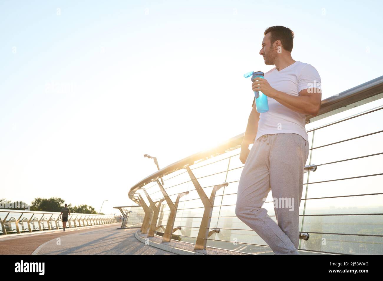 Full length portrait of a handsome sportsman athlete with a bottle of water, standing on the modern glass city bridge, relaxing after cardio workout o Stock Photo