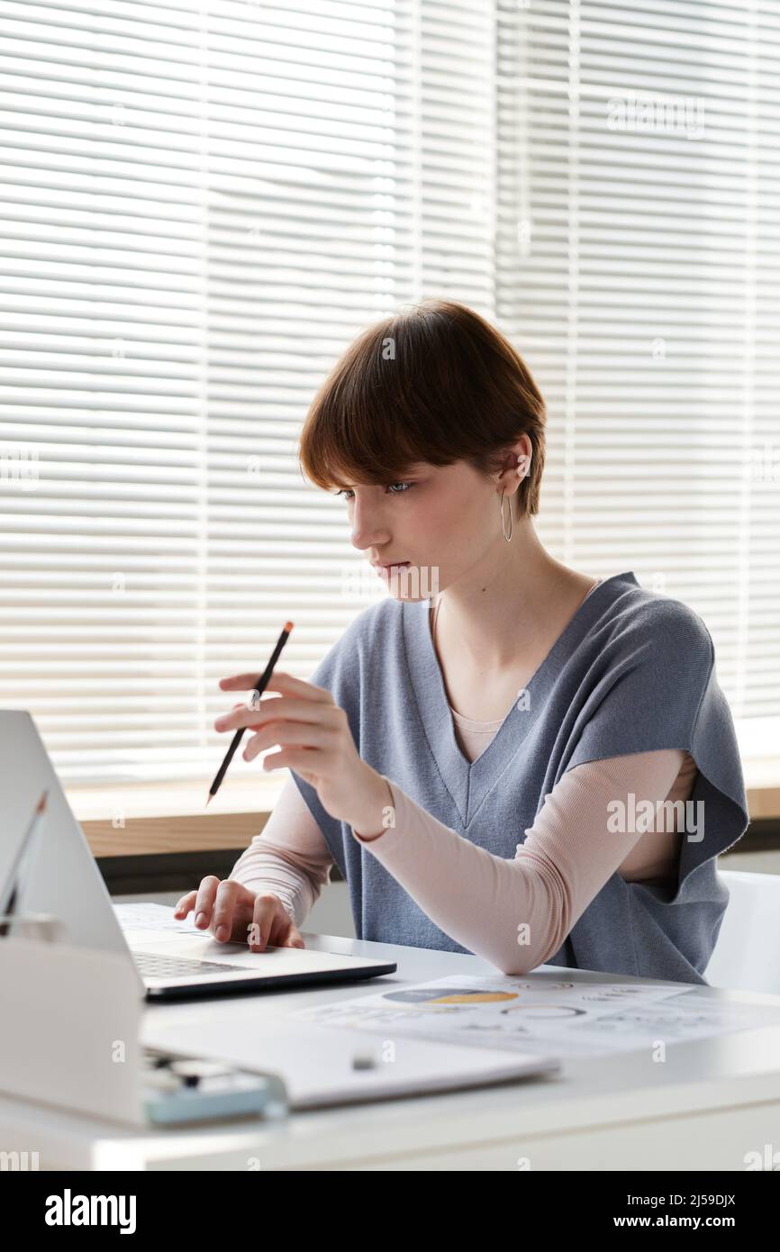 Pensive young businesswoman with short hair sitting at desk and twisting pencil while working with online data on laptop Stock Photo