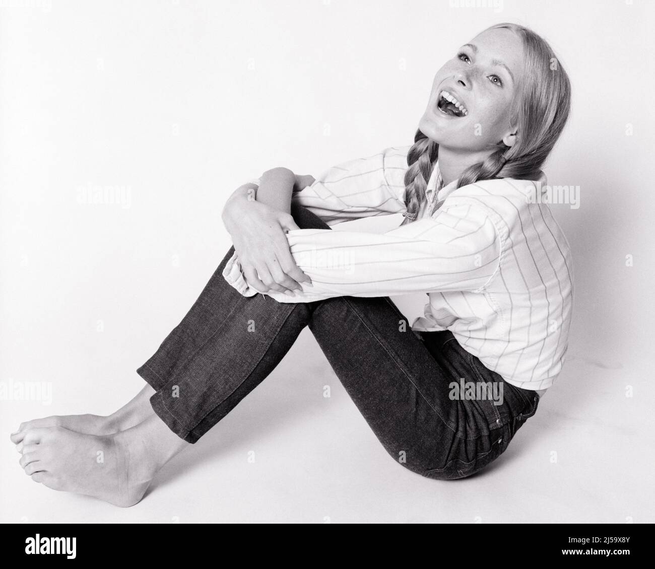 1960s LAUGHING TEEN GIRL SITTING ON FLOOR HUGGING HER KNEES LOOKING UP