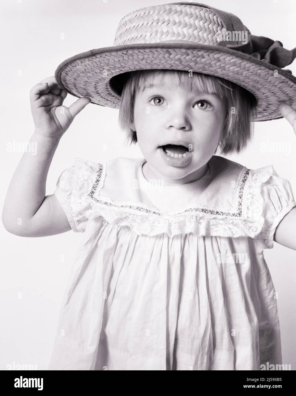 1960s CUTE LITTLE GIRL TODDLER HOLDING A TOO BIG STRAW HAT ON HER HEAD LOOKING AT CAMERA - j13195 HAR001 HARS STUDIO SHOT COPY SPACE EXPRESSIONS B&W EYE CONTACT WONDER HAPPINESS HEAD AND SHOULDERS DRESS UP STYLISH PLEASANT AGREEABLE CHARMING GROWTH JUVENILES LOVABLE PLEASING ADORABLE APPEALING BABY GIRL BLACK AND WHITE CAUCASIAN ETHNICITY HAR001 OLD FASHIONED Stock Photo