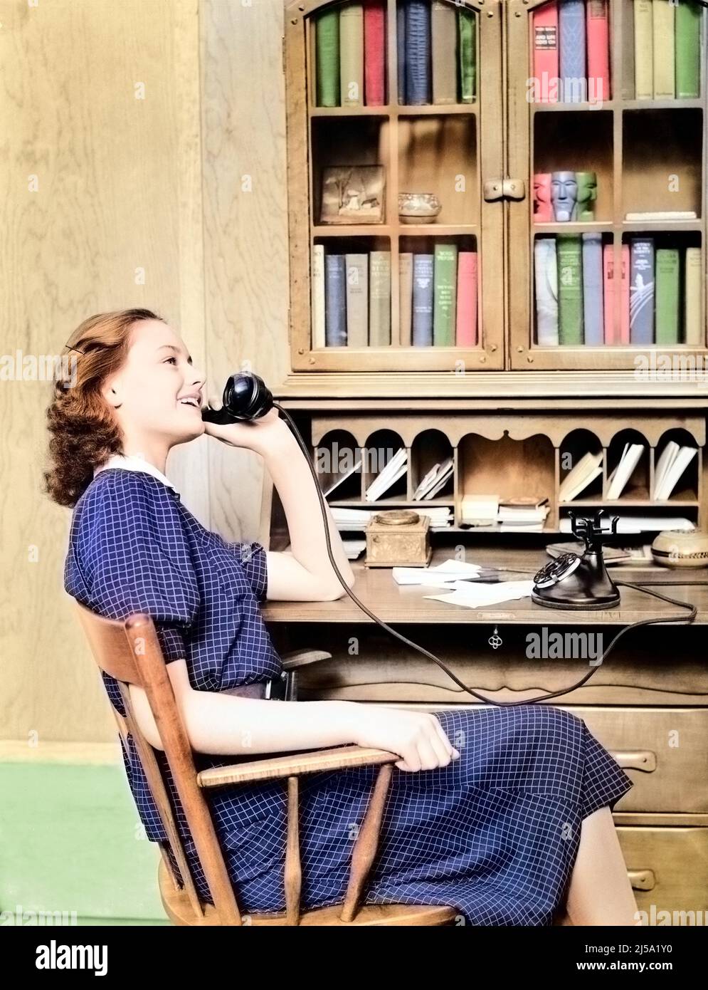 1930s 1940s TEENAGE GIRL SITTING AT DESK TALKING ON OLD BLACK ROTARY DIAL PHONE - t5280c HAR001 HARS STYLISH BOOKCASE ROTARY CAUCASIAN ETHNICITY HAR001 OLD FASHIONED Stock Photo