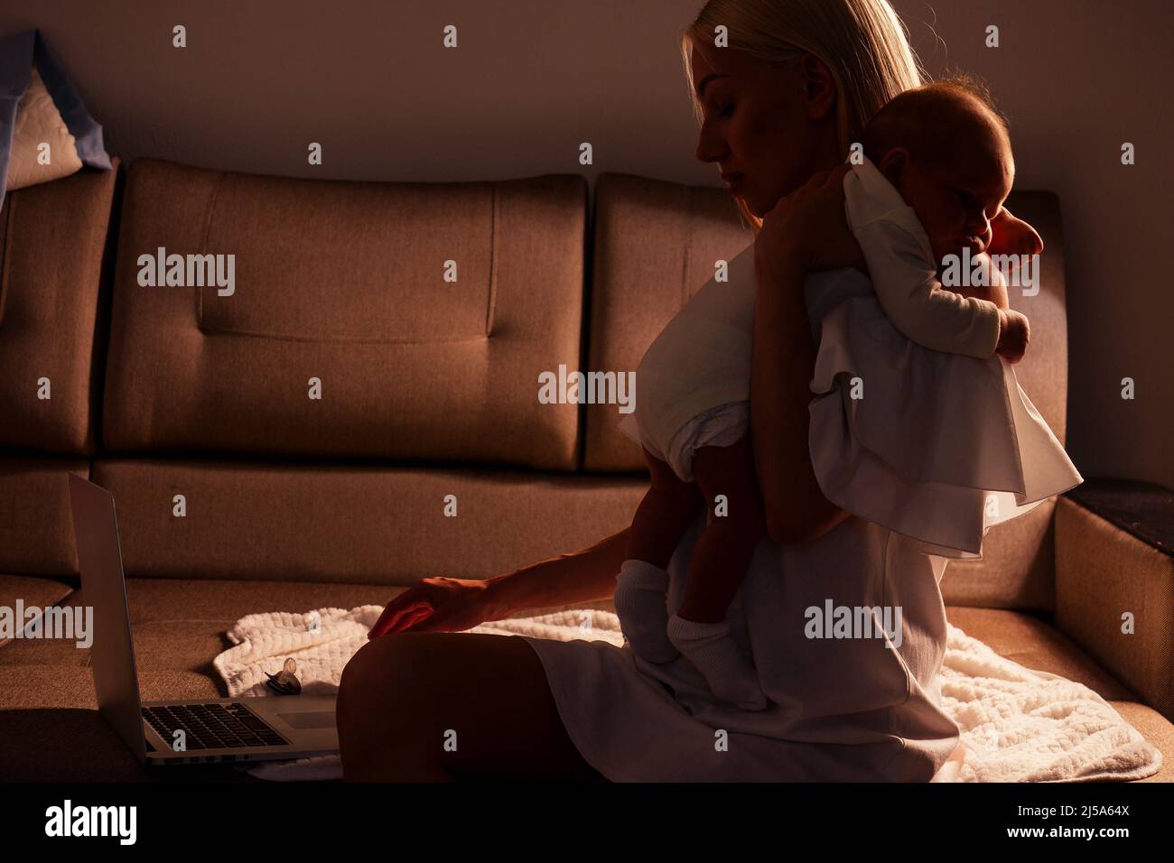 Happy single blonde mother working online and taking care of her baby surfing question on the internet in dark night room Stock Photo