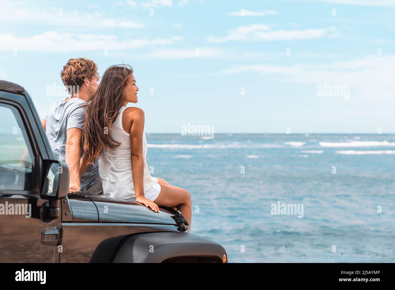 Car road trip travel couple tourists enjoying ocean view relaxing on hood of sports utility car. Happy Asian woman, man friends smiling on beach. Stock Photo