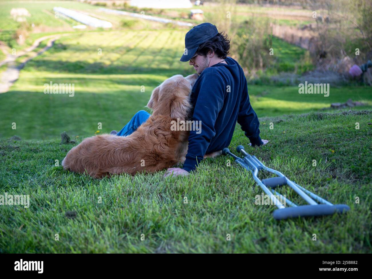 Crutches in the foreground of this idyllic rural farm background scene with a young man and his loving golden retriever dog cuddling with an arm aroun Stock Photo