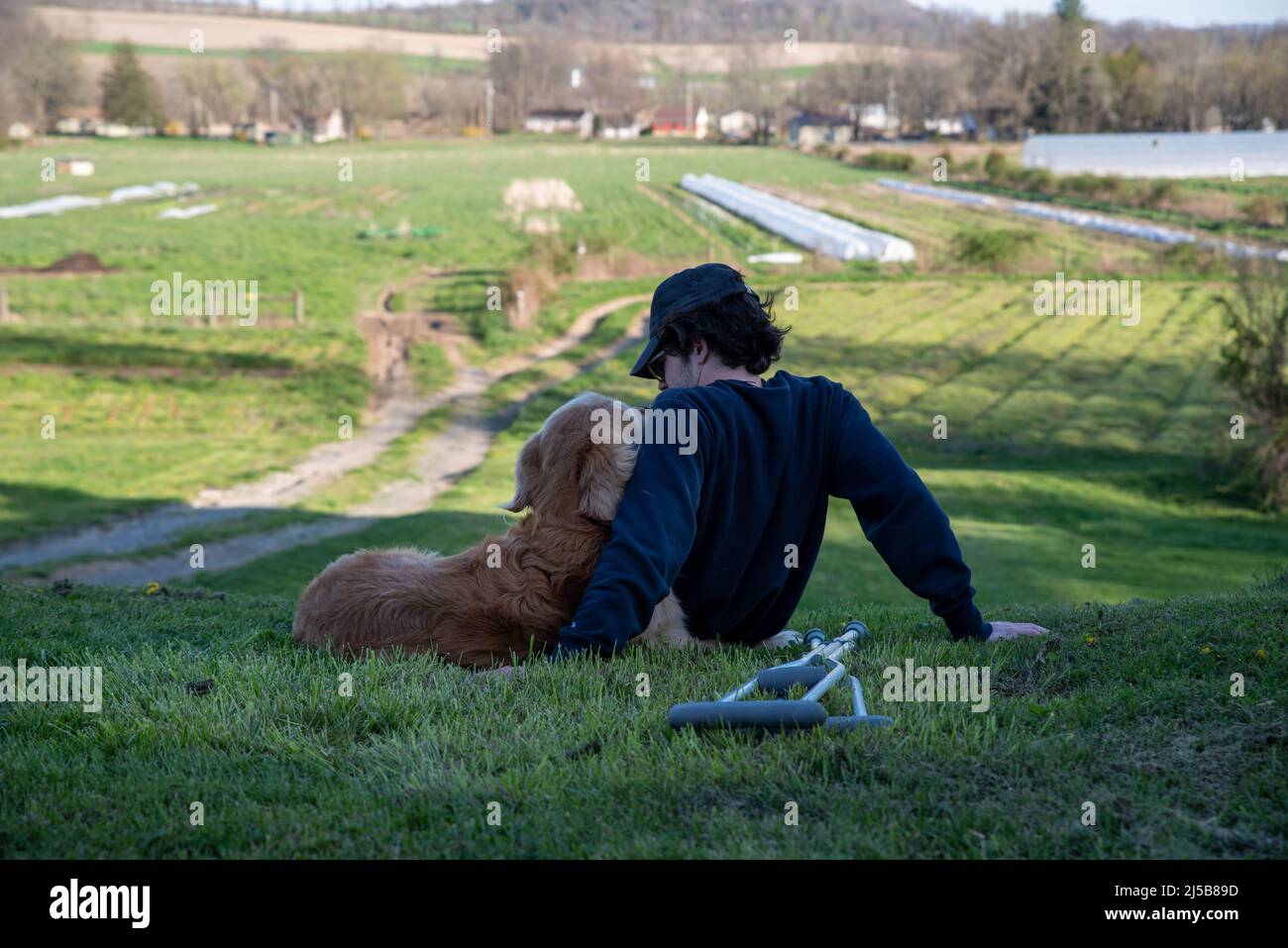 Crutches in the foreground of this idyllic rural farm background scene with a young man and his loving golden retriever dog cuddling and looking at ea Stock Photo
