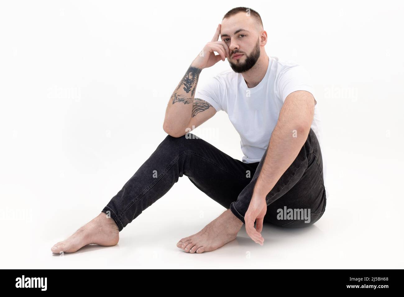 Young bearded man with short dark hair in white T-shirt, black jeans, sitting on white floor, leaning head on hand. Stock Photo