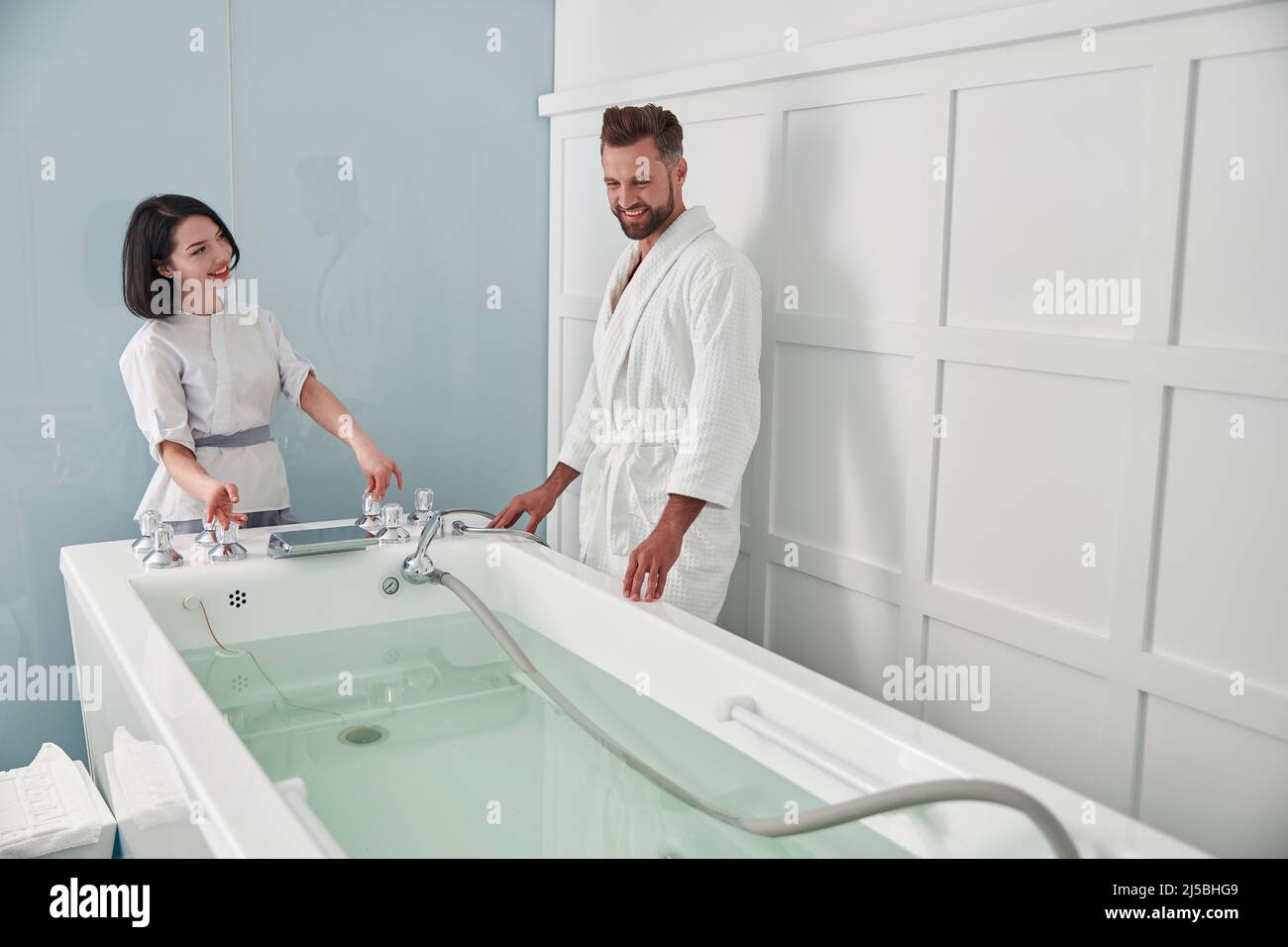 Specialist prepares hydro massage tub with clear water to mature guest in spa salon Stock Photo