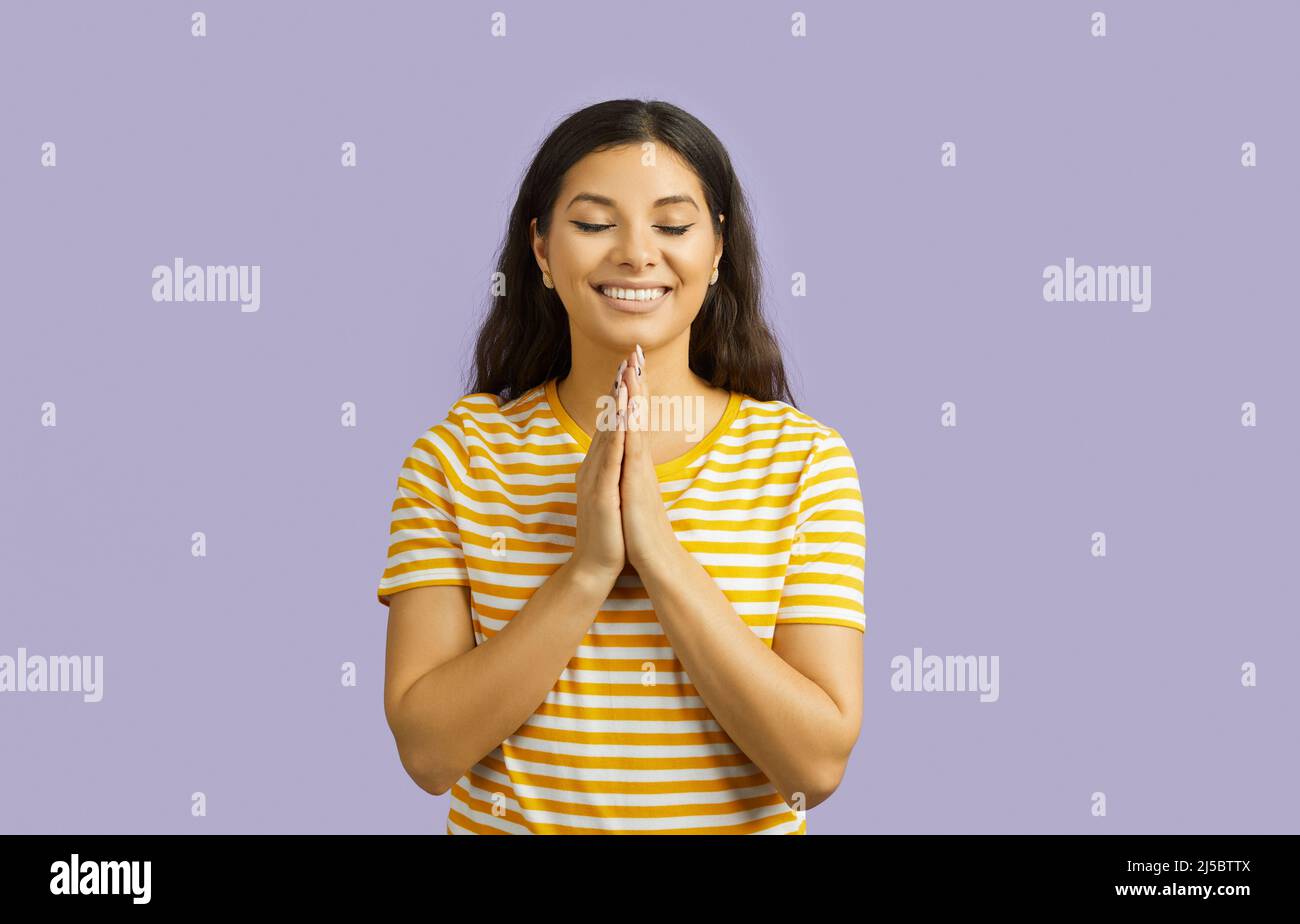Smiling young brunette woman t-shirt holding hands folded in prayer, keeping eyes closed posing isolated on pastel purple background. People lifestyle Stock Photo