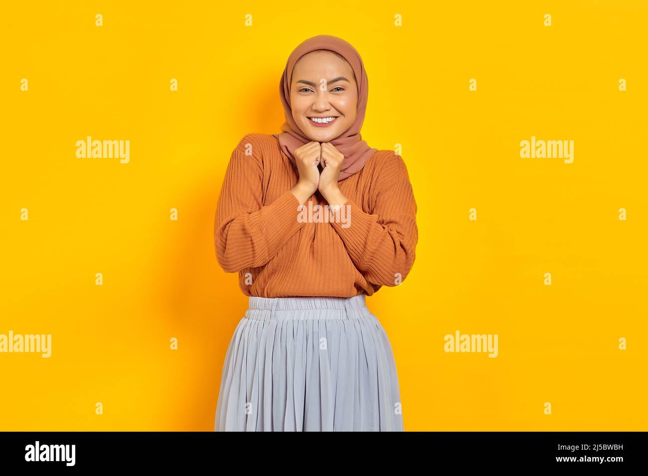 Beautiful smiling Asian woman in brown sweater and hijab keeps hands under chin, looking at camera, remembers pleasant moment isolated over yellow bac Stock Photo