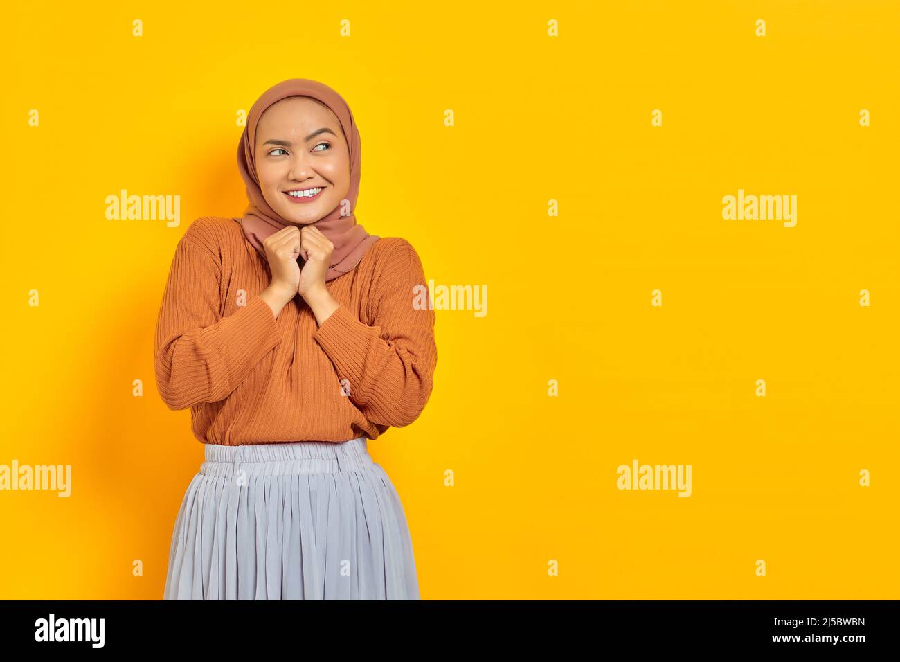 Beautiful smiling Asian woman in brown sweater and hijab keeps hands under chin, looking aside, remembers pleasant moment isolated over yellow backgro Stock Photo