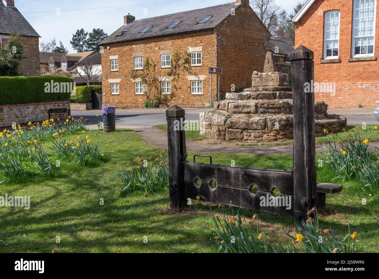 Remains of 16th century market cross with replica wooden stocks (from the 1960's), village green, Brixworth, Northamptonshire, UK Stock Photo