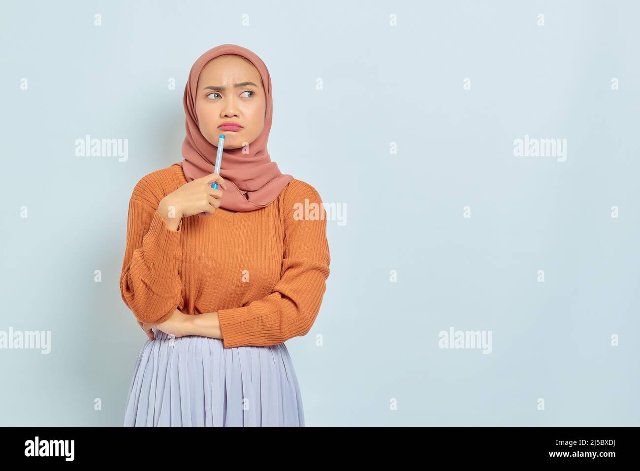 Shocked beautiful asian woman in brown sweater and hijab holding pen, looks serious thinking about a question isolated on white background. Muslim lif Stock Photo