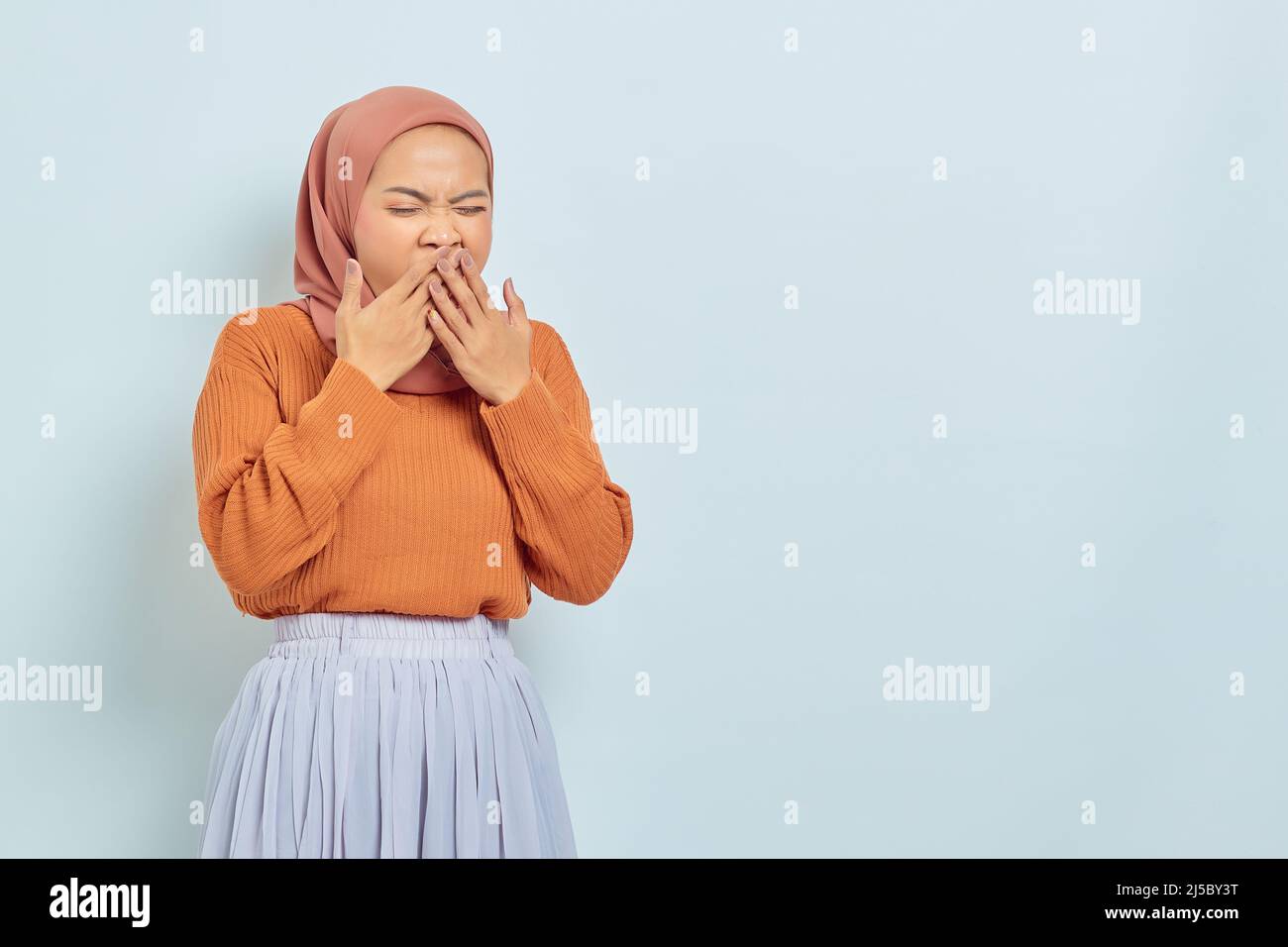 Beautiful Asian woman in brown sweater and hijab covering mouth and feeling sleepy isolated on white background. Stock Photo