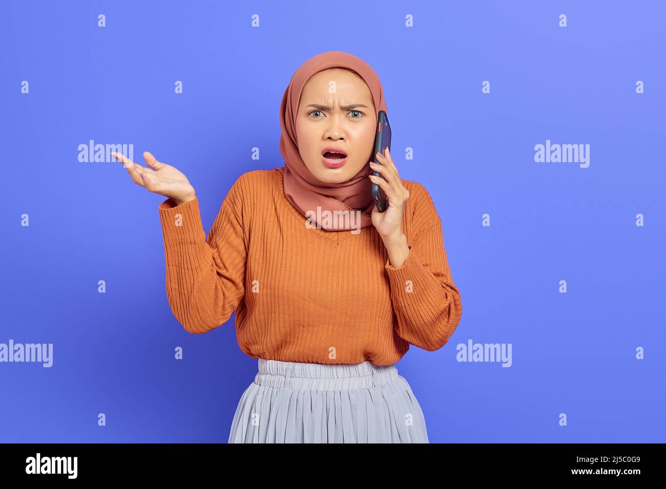 Shocked beautiful Asian woman in brown sweater and hijab talking on