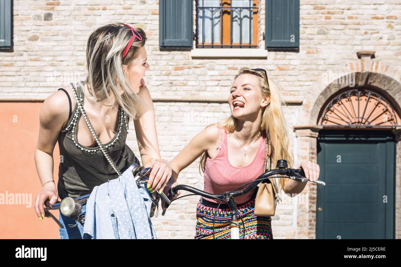 Happy female friends couple having fun riding bicycle in city old town - Friendship concept with young girlfriend on funny attitude biking together in Stock Photo