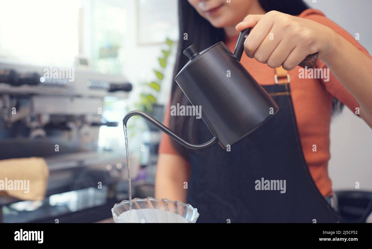 A female café operator wearing an apron pours hot water over roasted coffee grounds to prepare coffee for customers in the shop. Stock Photo