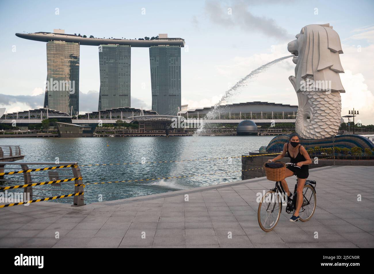 28.05.2020, Singapore, Republic of Singapore, Asia - A woman rides her bicycle along the otherwise deserted Merlion Park on the banks of the Singapore River during curfew, wearing a mouth guard to protect herself from contracting the pandemic coronavirus. In the background you can see the Marina Bay Sands Hotel. Usually the area around Marina Bay is teeming with tourists, but now it is completely deserted except for a few people due to Covid-19. [automated translation] Stock Photo