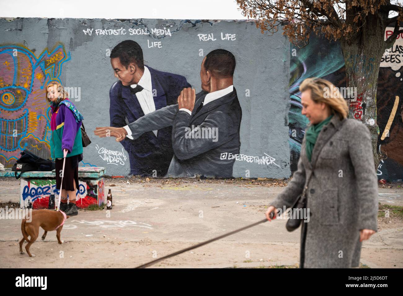 '30.03.2022, Berlin, Germany, Europe - Two women walk their dogs while in the background graffiti by artist Eme Freethinker is seen on a segment of the Berlin Wall in Berlin's Mauerpark in the Prenzlauer Berg district, showing actor Will Smith slapping comedian Chris Rock on stage at the 94th Academy Awards after he made fun of his wife Jada Pinkett Smith. Will Smith later won the first Oscar in his career for the role of the father of tennis players Venus and Serena Williams in the movie ''King Richard''. [automated translation]' Stock Photo