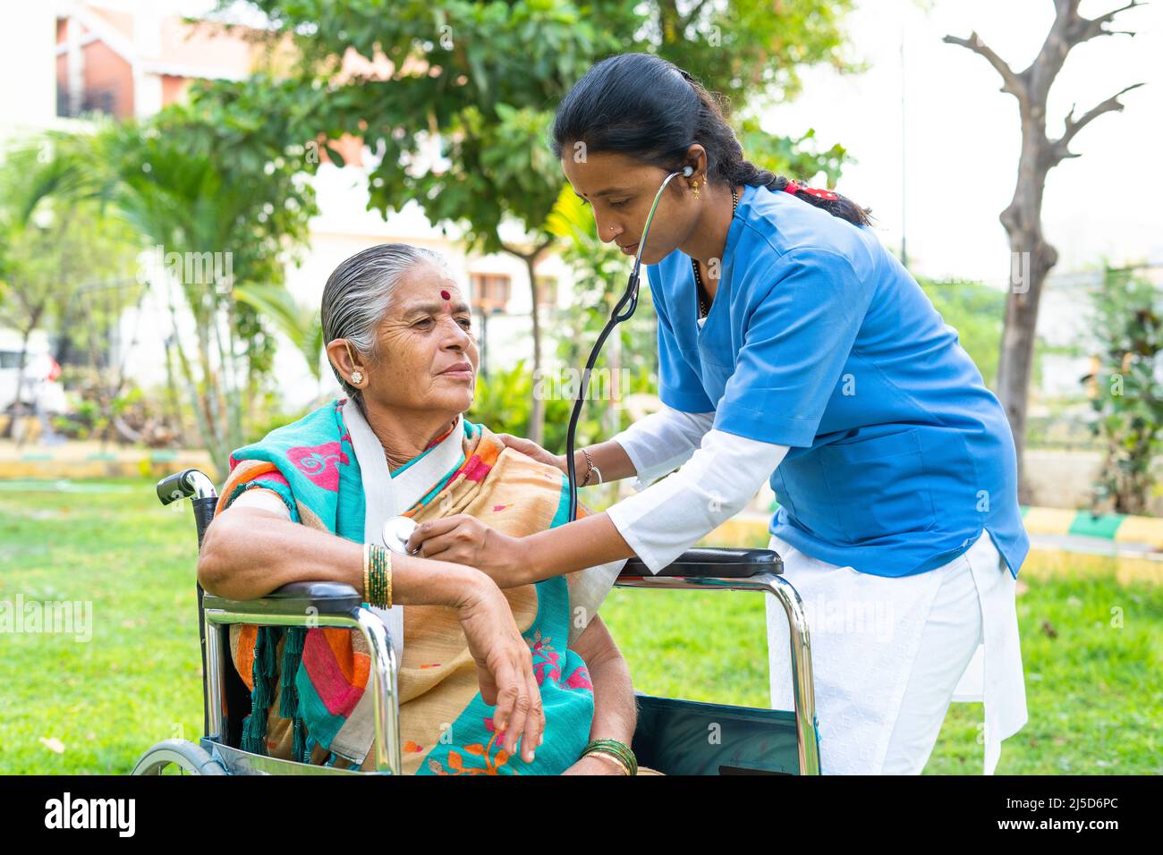Nurse busy checking or monitoring health of senior woman with disability on wheelchair at hospital park - concept of healthcare, professional Stock Photo