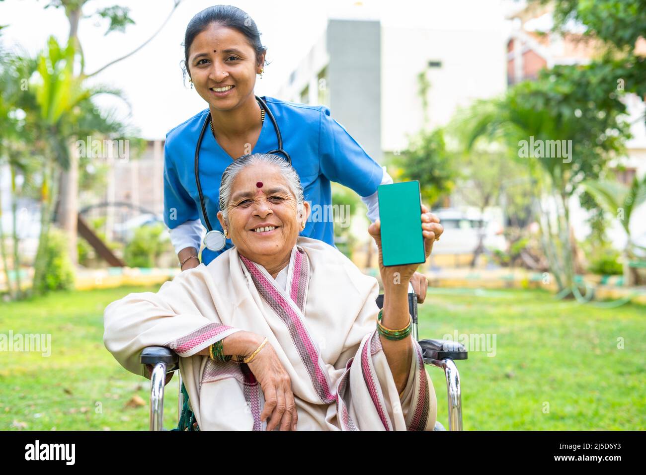 Senior women showing green screen mobile phone while with nurse by looking at camera at hospital park - concept of online Healthcare booking service Stock Photo