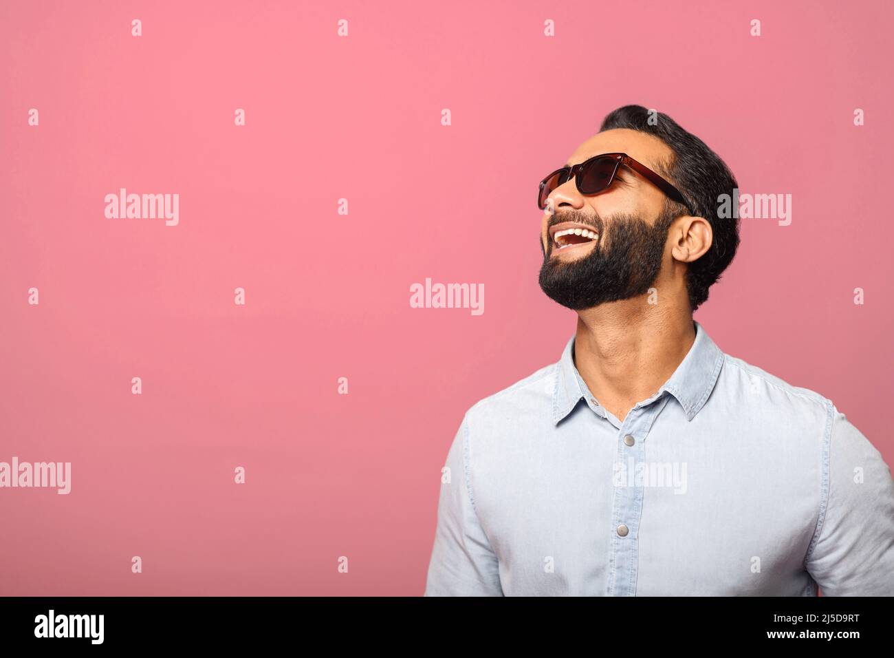 Cheerful Indian man in sunglasses laughing toothy, carefree hispanic guy in blue shirt standing and laughs out loud over pink background. Happy lifestyle Stock Photo