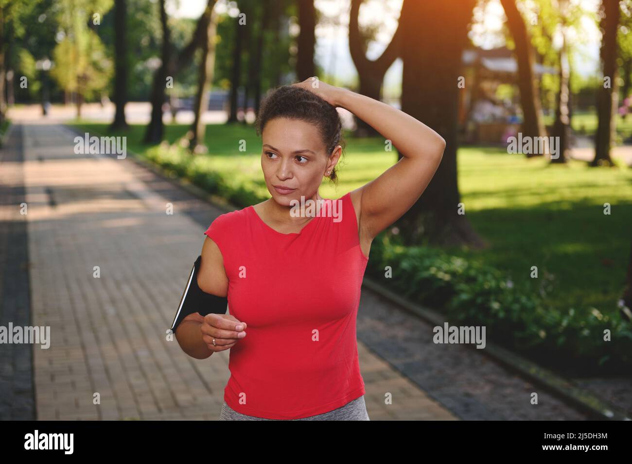Determined middle-aged sportswoman in red t-shirt with smartphone holder tying ponytail while standing on path during morning jog along park. Cardio w Stock Photo