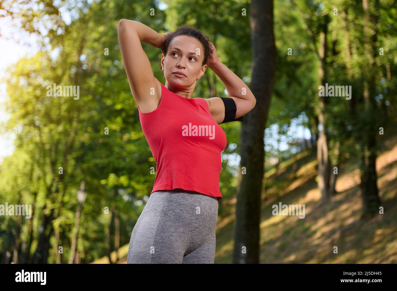 Confident portrait of a beautiful fit woman in sportswear with a smartphone holder tying a ponytail, standing in a city park on a sunny warm summer da Stock Photo