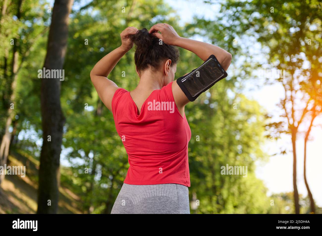 Rear view of a young determined sportswoman wearing a smartphone holder, tying ponytail, getting ready for run along the city park outdoor Stock Photo