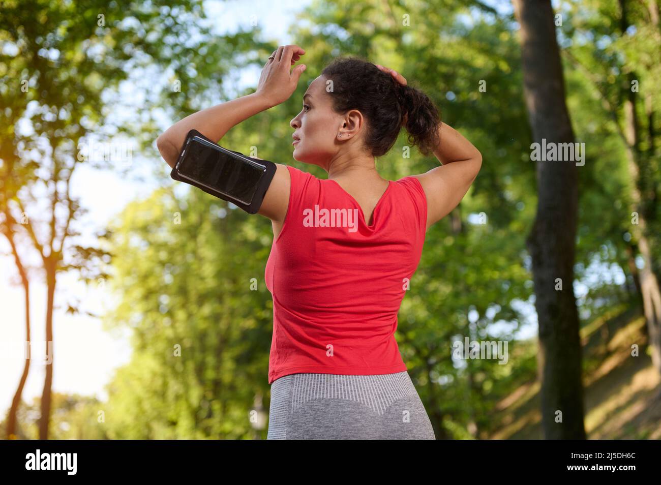 Rear view of a sportswoman wearing a smartphone holder, tying ponytail, getting ready for run along the city park outdoor Stock Photo