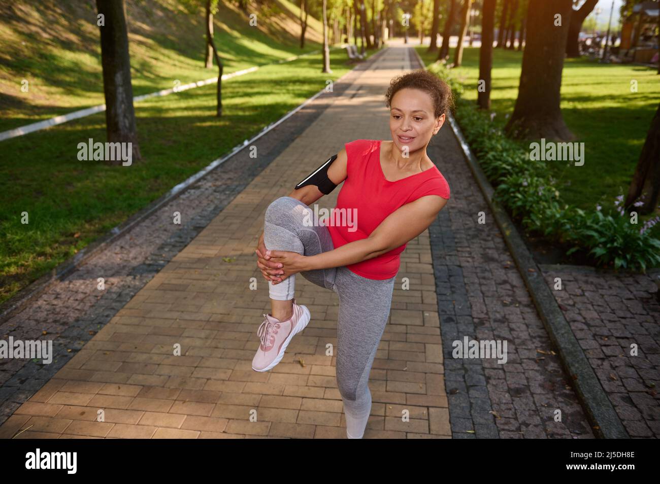 Determined cheerful athlete, fit woman in gray leggins and red tight t-shirt exercising outdoor in the city park. Fitness, body weight training outdoo Stock Photo