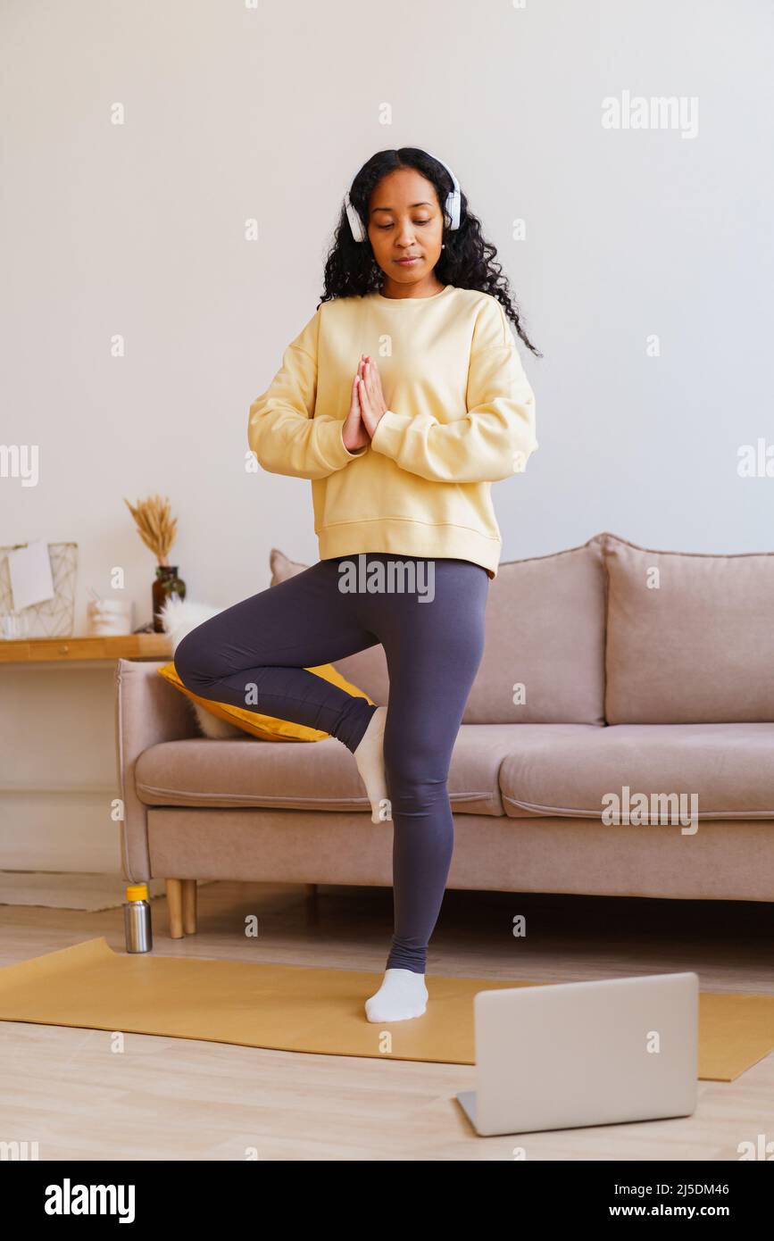 African-American female in headphones standing in tree pose yoga while following tutorial on laptop Stock Photo