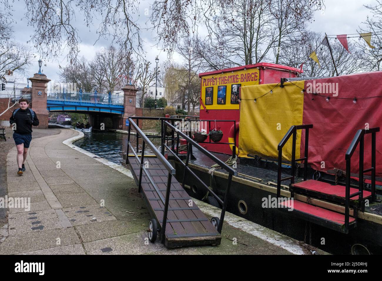Man walks along Regent’s Canal tow path at Blomfield Road, Little Venice next to Puppet Theatre Barge. Warwick Avenue in background. West London, UK. Stock Photo