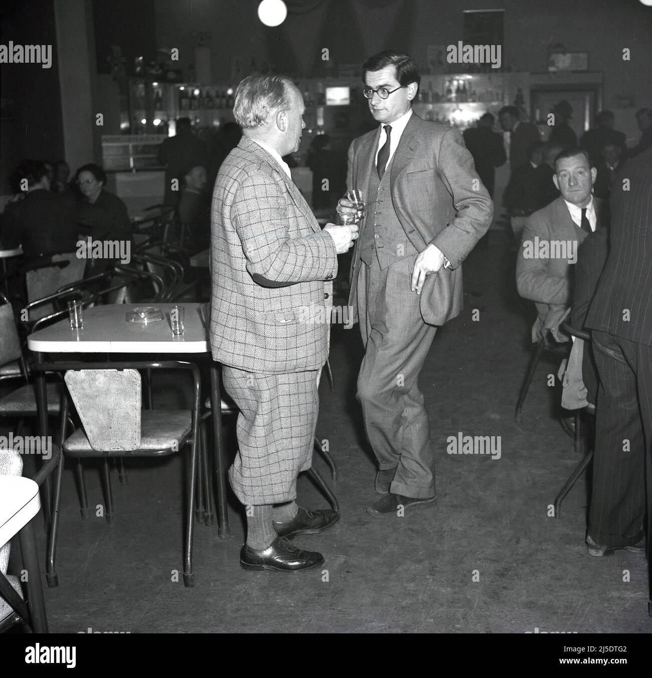 1950s, historical, two well-dressed men, talking together with a drink in hand. One is in a check suit with plus fours, the other in a suit & tie and waistcoat and they are standing chatting inside the social club of the Steel Company of Wales at the Abbey steelworks in Port Talbot, Wales, UK. Stock Photo